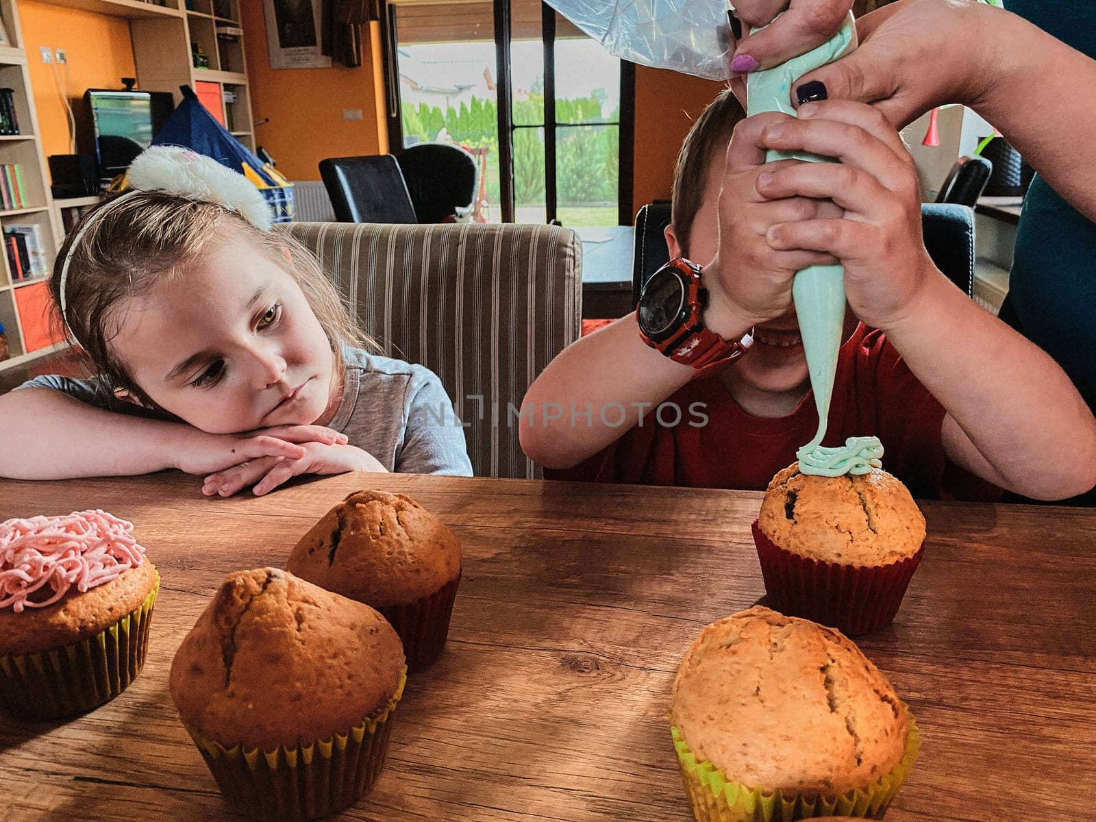 Group of children baking cupcakes, squeezing cream from confectionery bag, preparing ingredients, topping, sprinkles for decorating cookies. Kids cooking, working together in kitchen at home. Concept of happy family