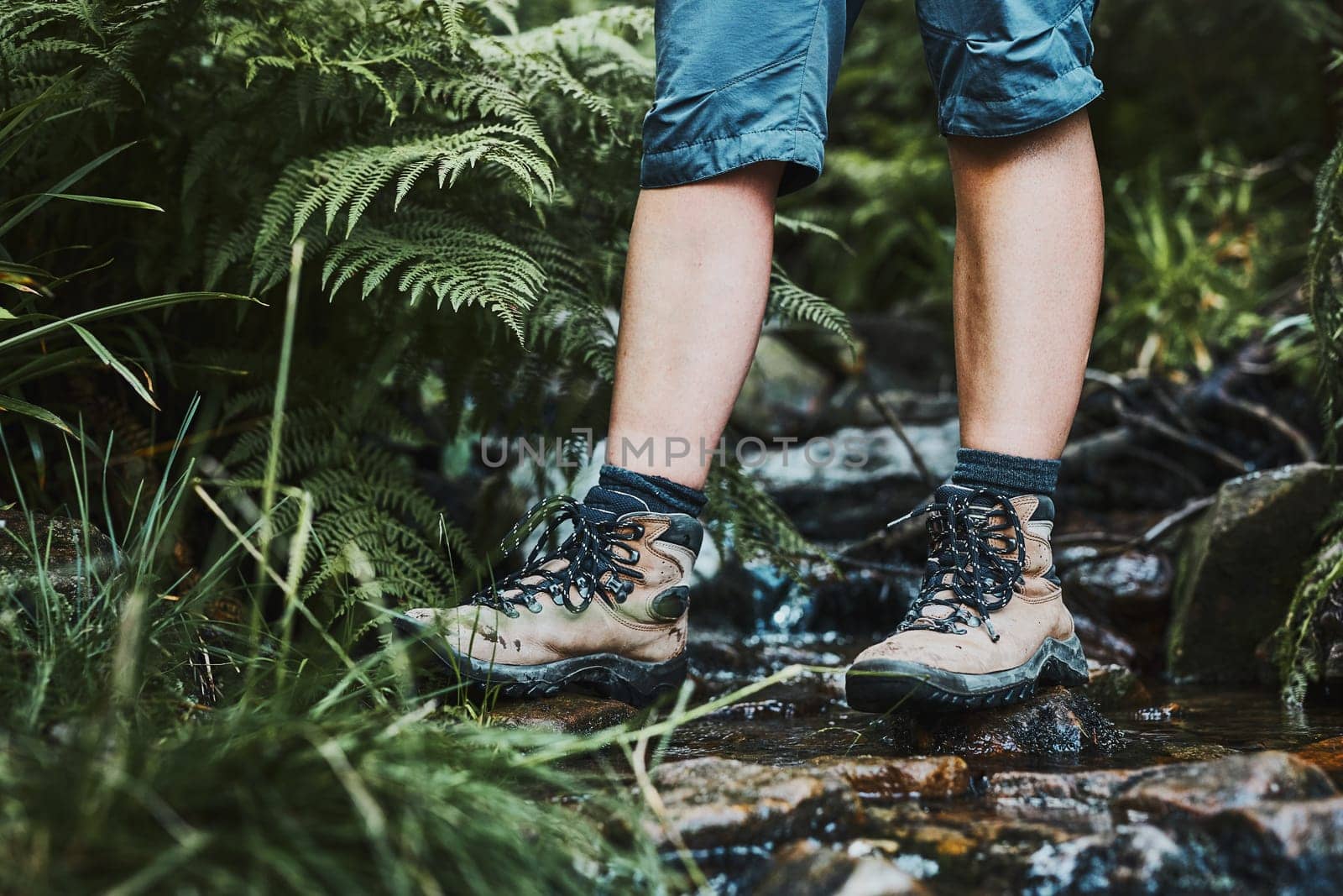 Female legs with trekking boots standing on stones in mountain stream. Summer vacation close to nature. Concept of actively spending leisure time