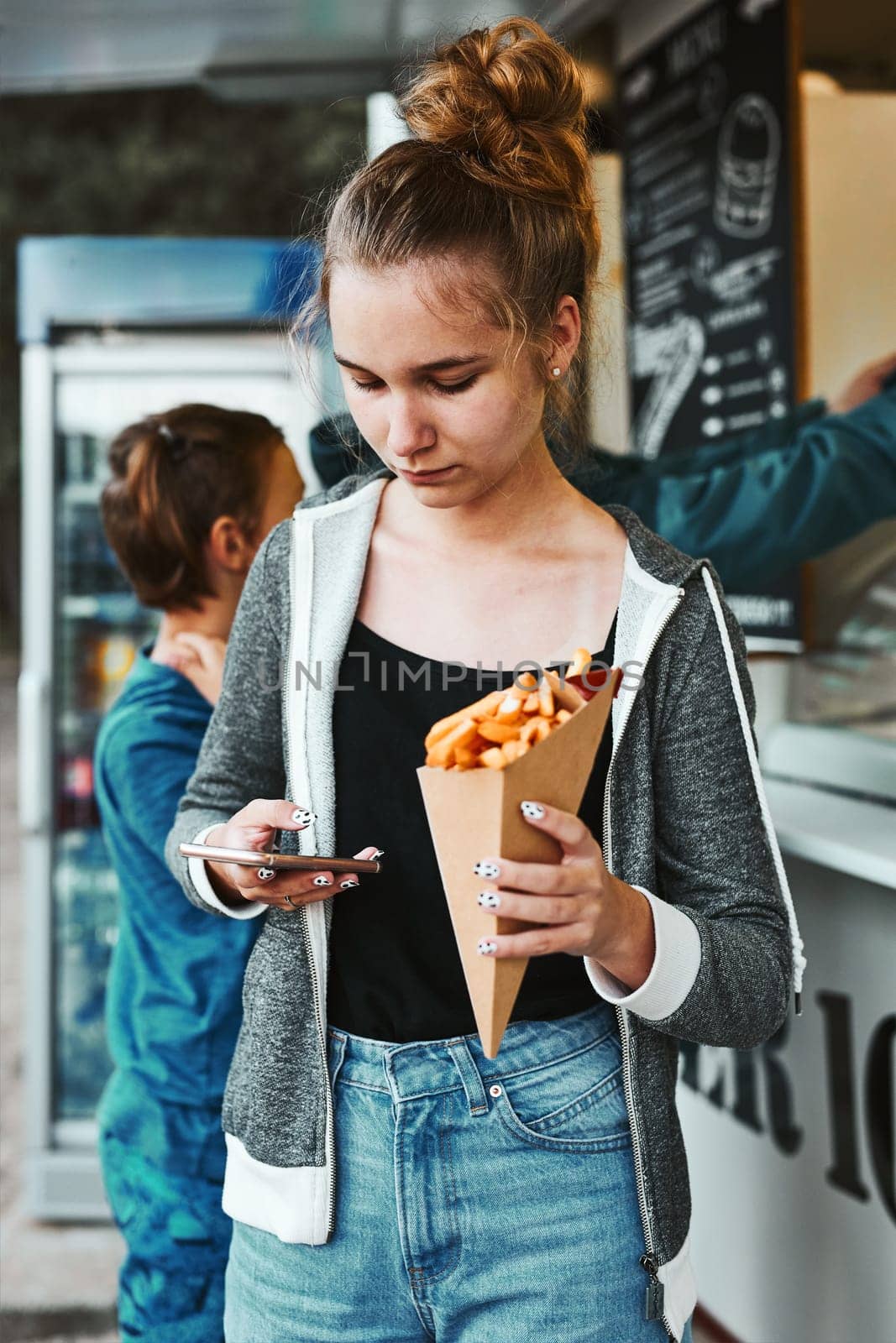 Teenage girl eating potato fries from carton cone standing in front of food truck. Teenager having a fast food meal outdoors during summer vacations