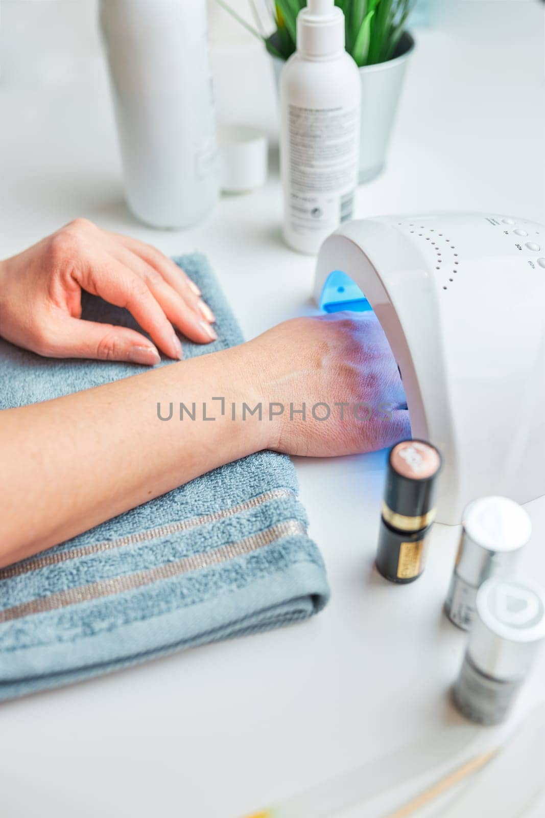 Woman applying gel hybrid polish using UV lamp. Beauty wellness spa treatment concept. Cosmetic products, UV lamp, green leaves on white table. Spa, manicure, skin care concept. Flat lay, overhead view