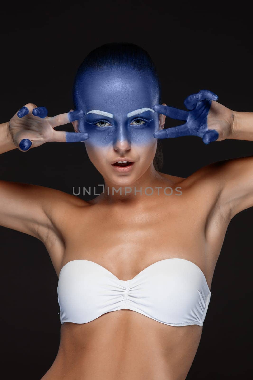 Portrait of a young woman who is posing covered with blue paint in the studio on a black background. girl&#39 s hands around the face and painted blue