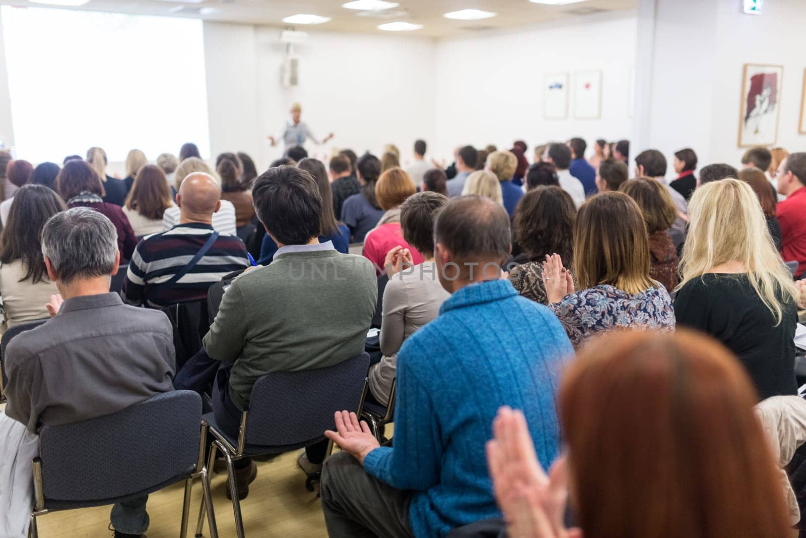 Life coaching symposium. Female speaker giving interactive motivational speech at entrepreneurship workshop. Audience in conference hall. Rear view of unrecognized participant.