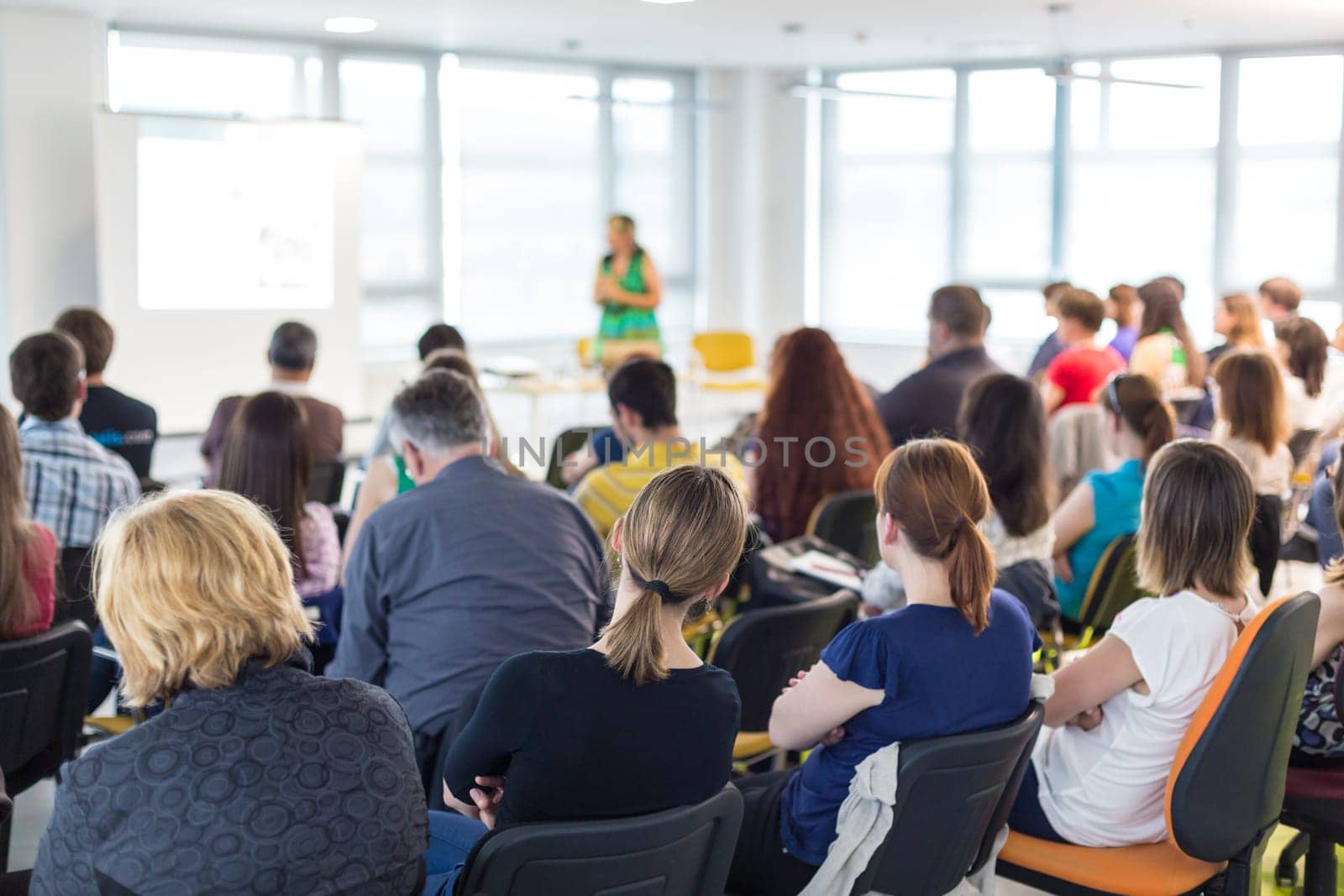 Business and entrepreneurship symposium. Female speaker giving a talk at business meeting. Audience in conference hall. Rear view of unrecognized participant in audience.