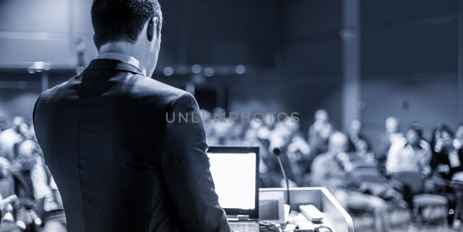 Speaker giving a talk on corporate business conference. Unrecognizable people in audience at conference hall. Business and Entrepreneurship event. Blue toned grayscale image.