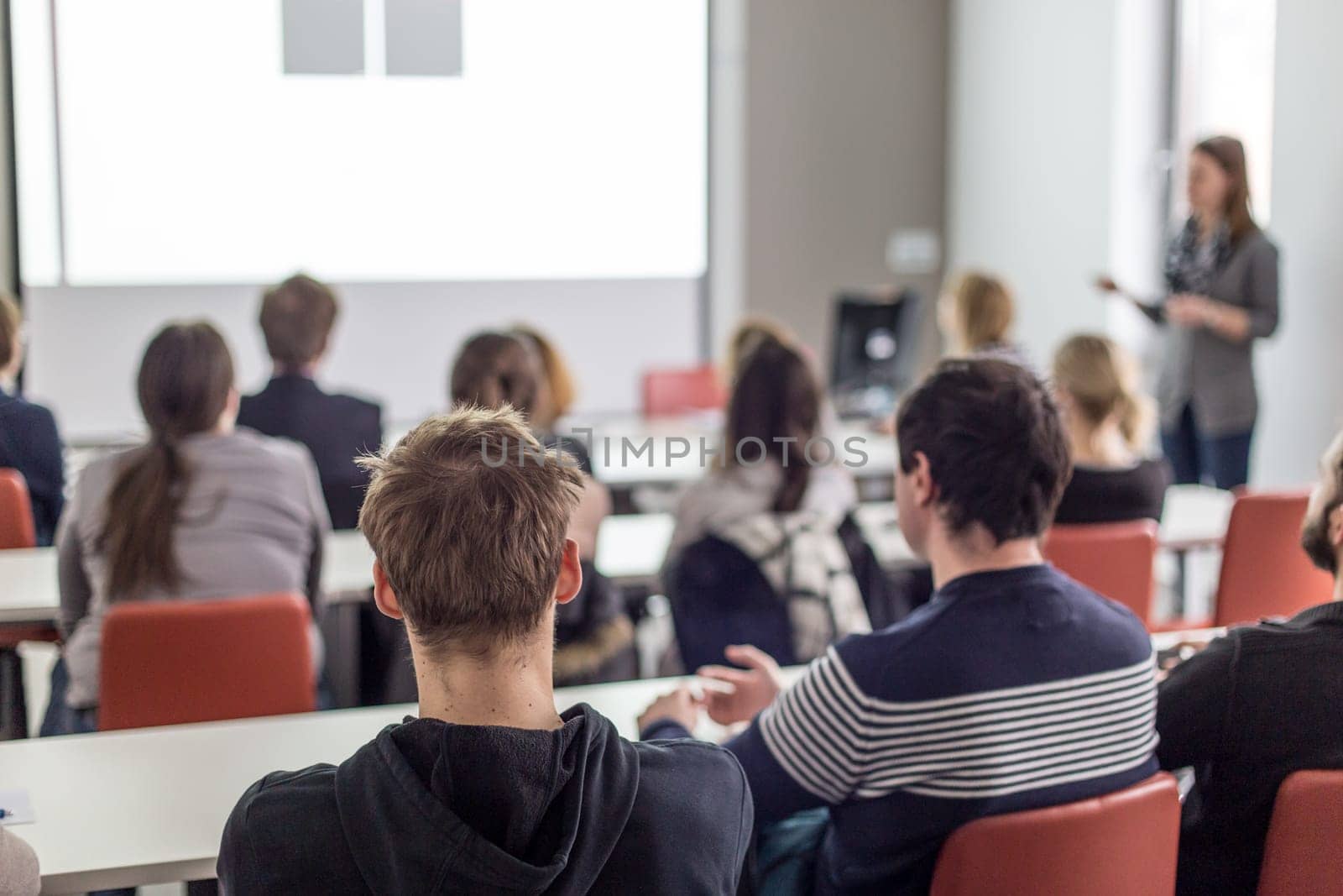 Female speaker giving presentation in lecture hall at university workshop. Audience in conference hall. Rear view of unrecognized participant in audience. Scientific conference event.