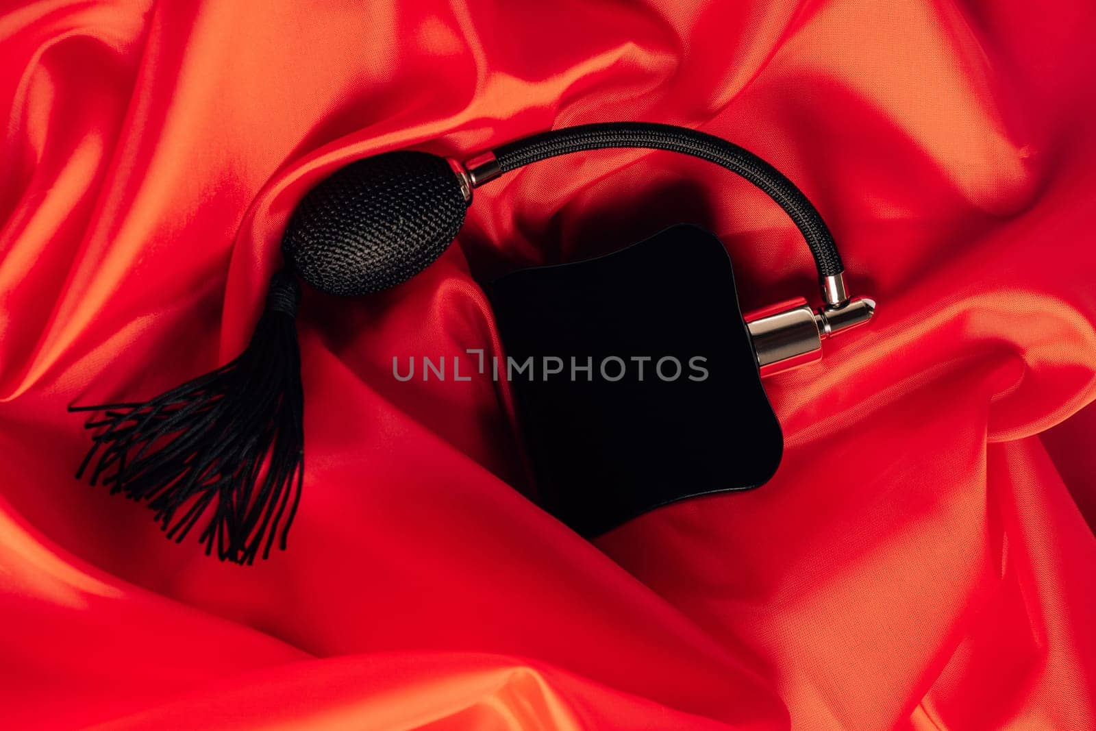 Black bottle of eau de toilette or perfume with long tassel spray pomp lies on red background of flowing fabric