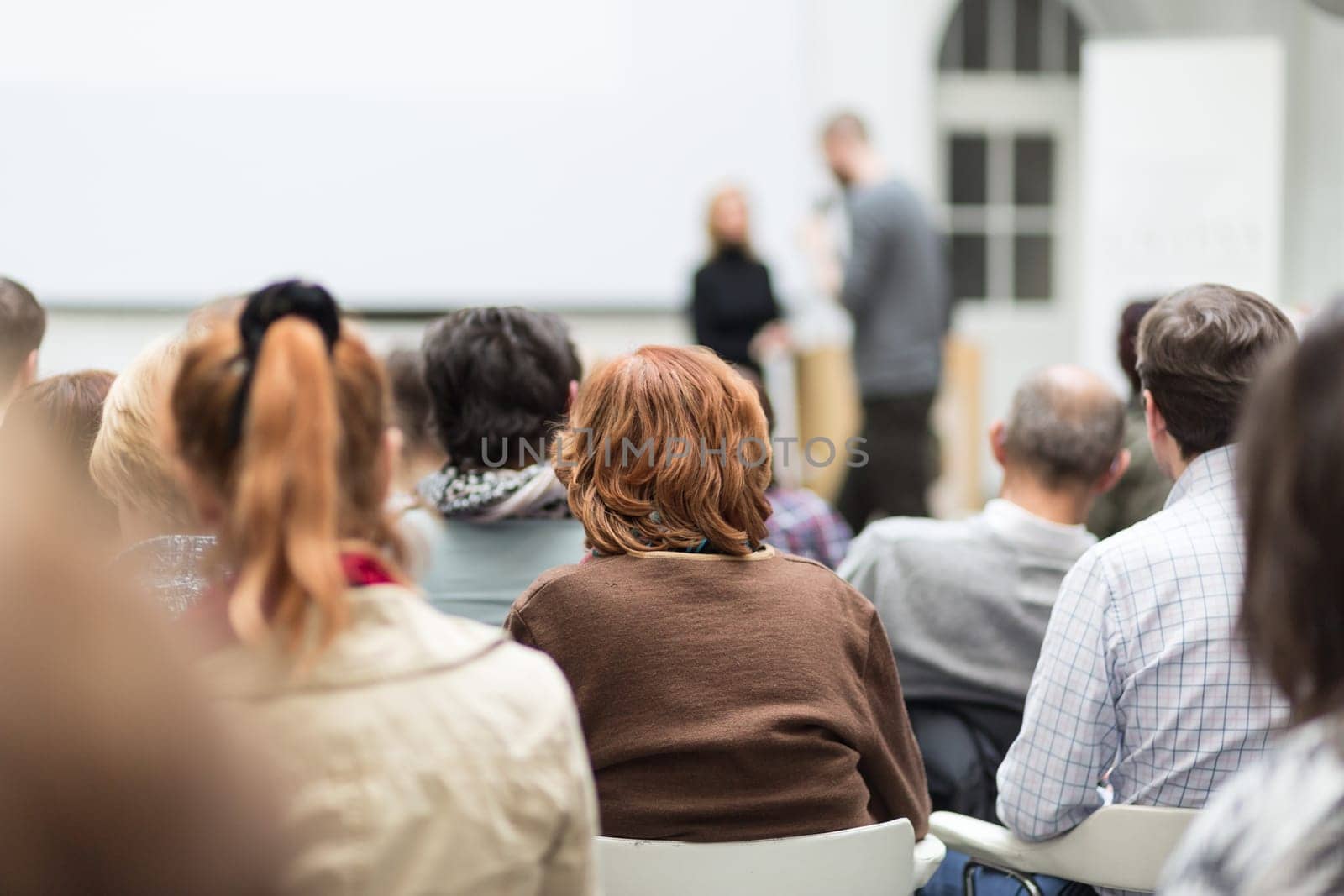 Business and entrepreneurship symposium. Female speaker giving a talk at business meeting. Audience in conference hall. Rear view of unrecognized participant in audience. Copy space on whitescreen.
