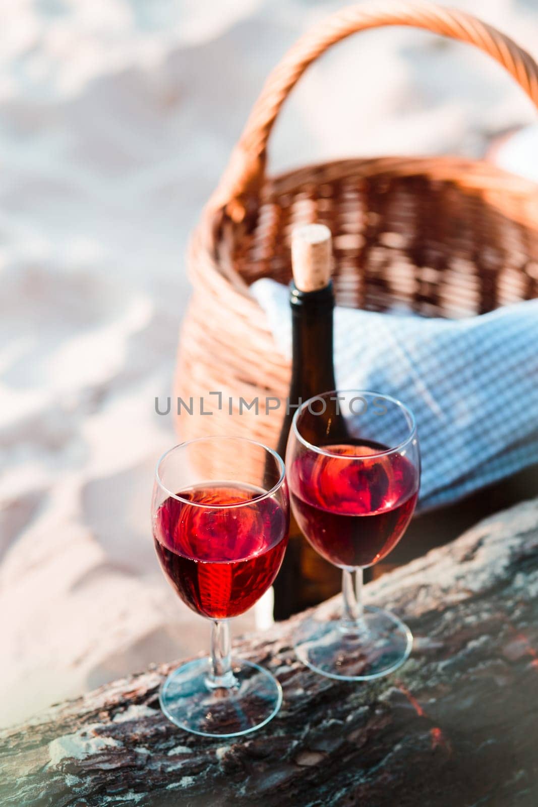 Two wine glasses with red wine standing on tree trunk, on beach, beside wicker basket with bottle of wine