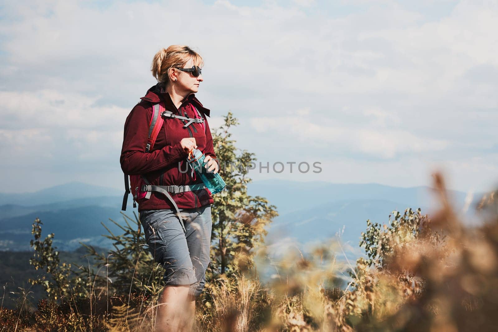 Woman with backpack hiking in mountains, spending summer vacation close to nature. Woman walking on top of hill admiring mountain landscape panorama holding bottle with water