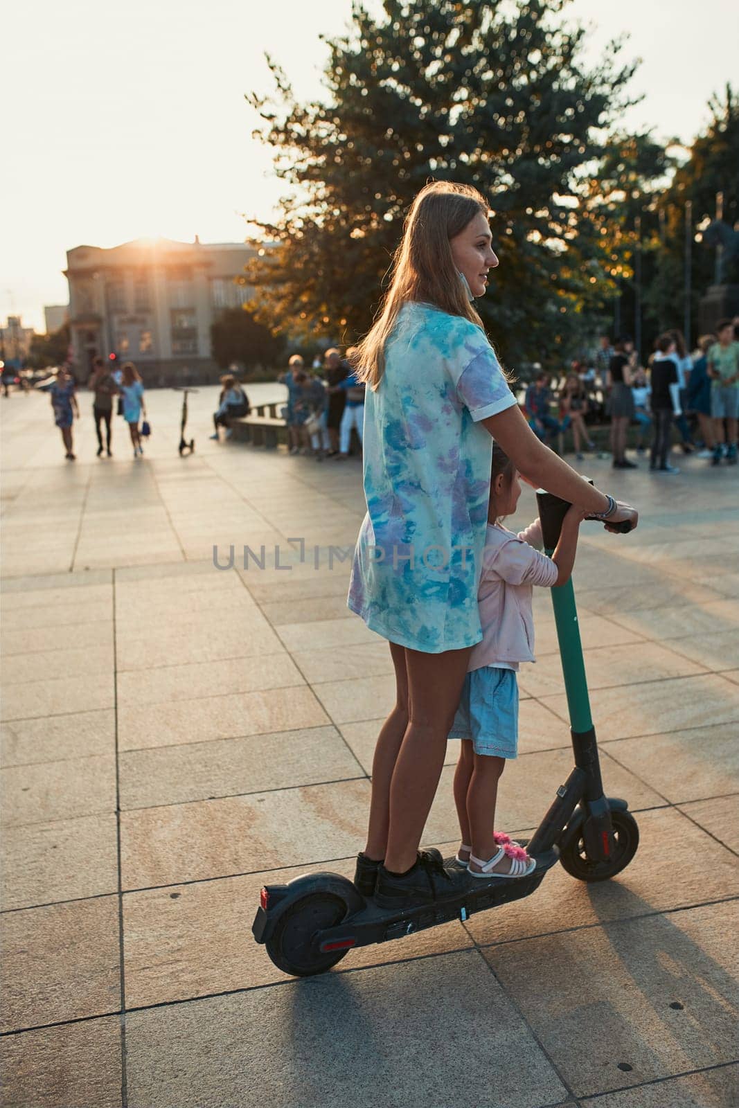 Teenager girl and her sister preschooler riding an electric scooter in the city center. Concept of happy family. Candid people, real moments, authentic situation
