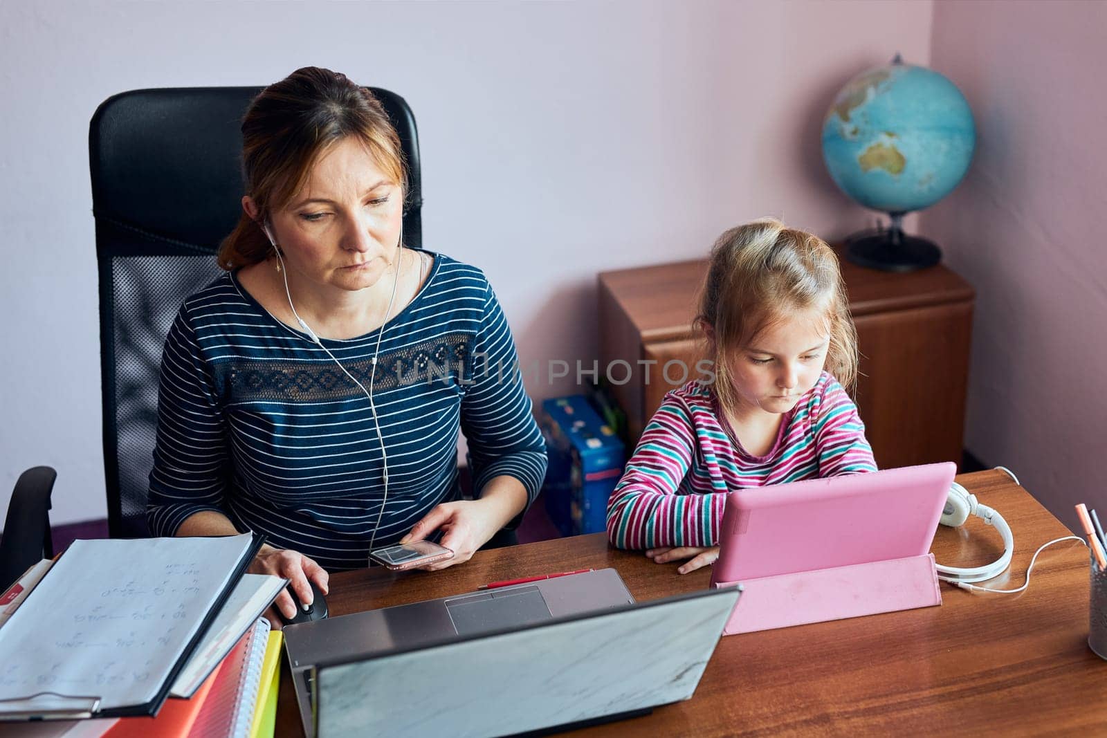 Woman mother working doing her job remotely during video chat call stream online course webinar on laptop from home while her daughter watching video on tablet. Woman sitting at desk in front of computer looking at screen