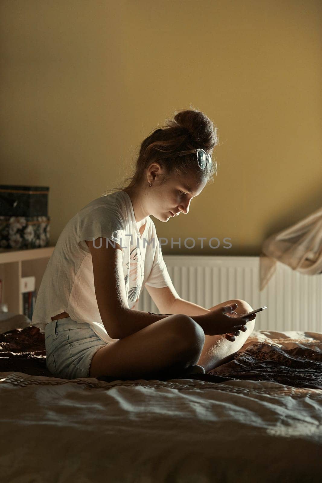 Young woman using portable computer and mobile phone, sitting on a bed, learning online at home. Candid people, real moments, authentic situations