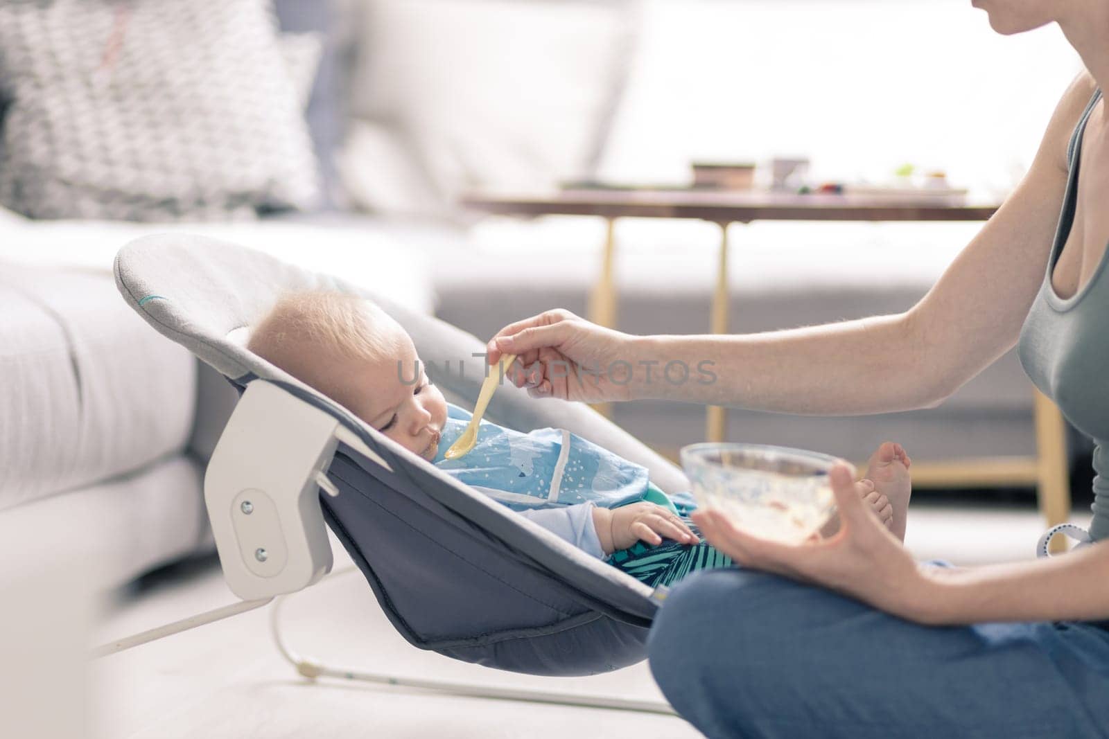 Mother spoon feeding her baby boy child in baby chair with fruit puree. Baby solid food introduction concept