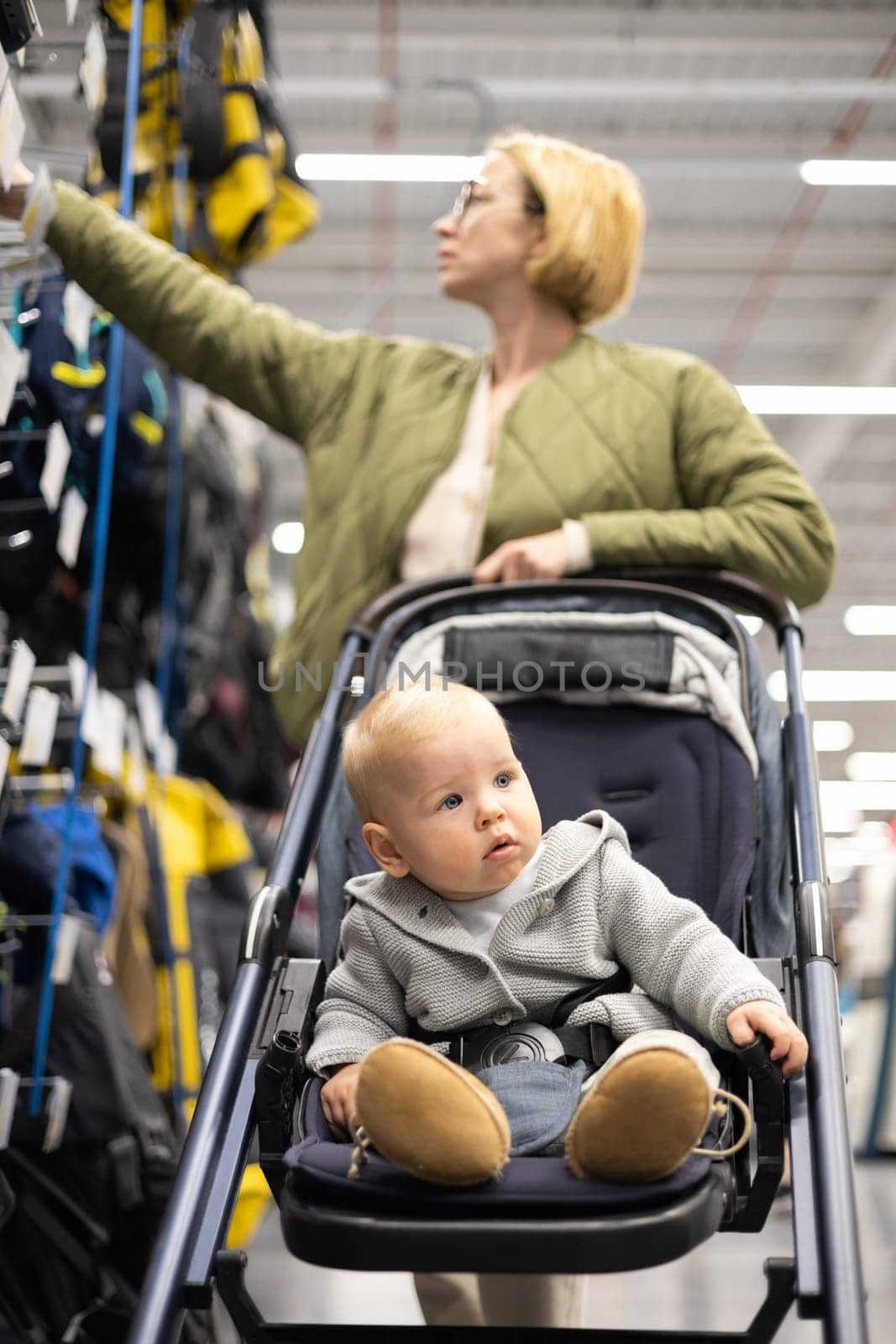 Casualy dressed mother choosing sporty shoes and clothes products in sports department of supermarket store with her infant baby boy child in stroller