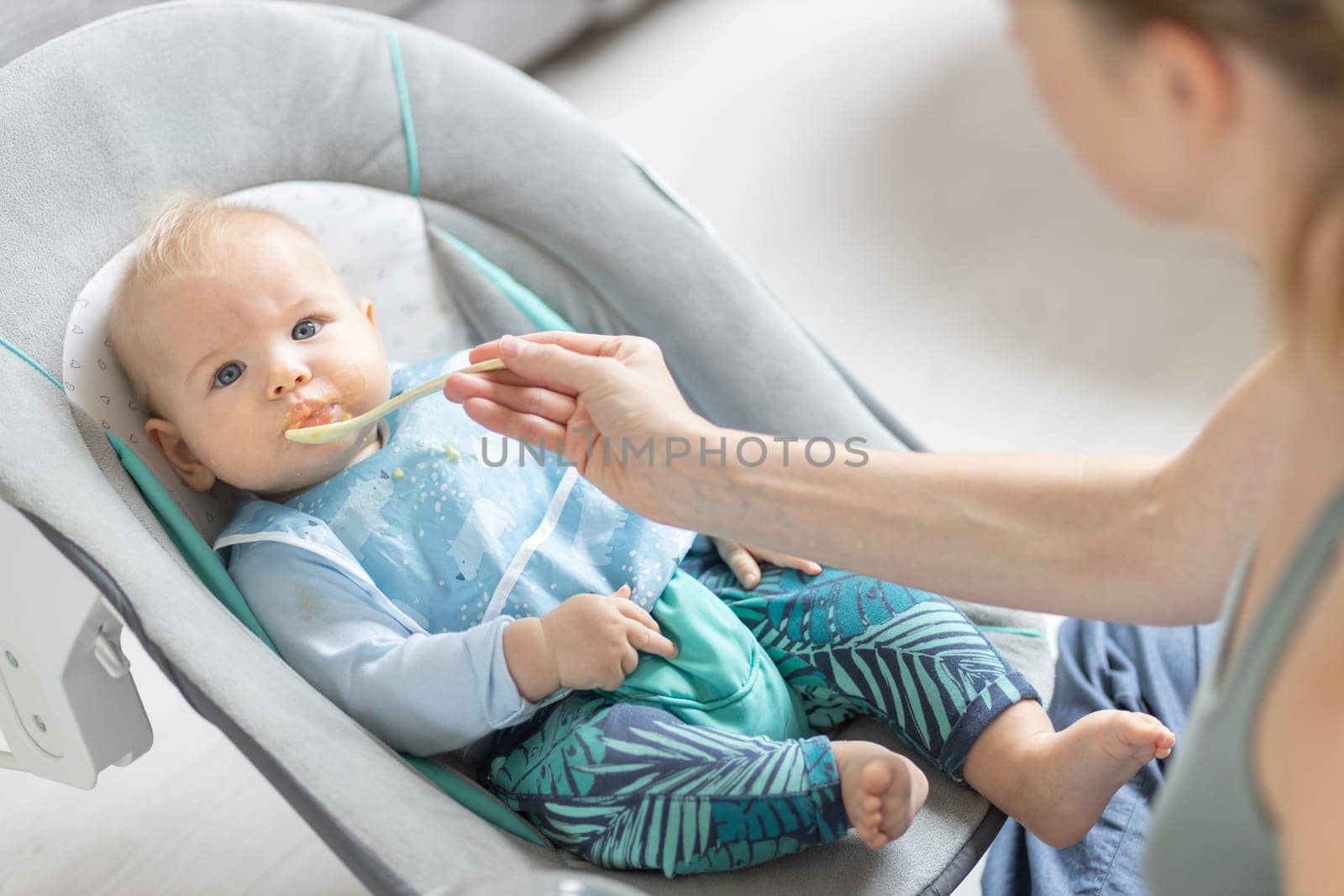 Mother spoon feeding her baby boy child in baby chair with fruit puree. Baby solid food introduction concept