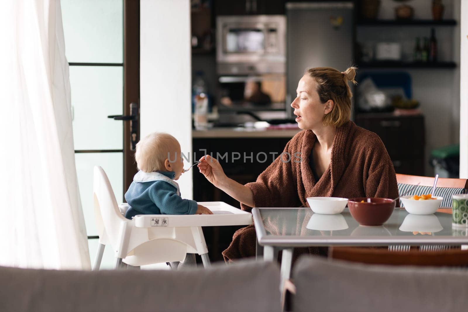 Mother wearing bathrope spoon feeding her infant baby boy child sitting in high chair at the dining table in kitchen at home in the morning.
