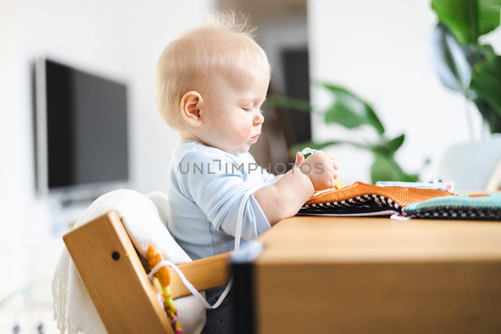 Happy infant sitting at dining table and playing with his toy in traditional scandinavian designer wooden high chair in modern bright atic home. Cute baby playing with toys.