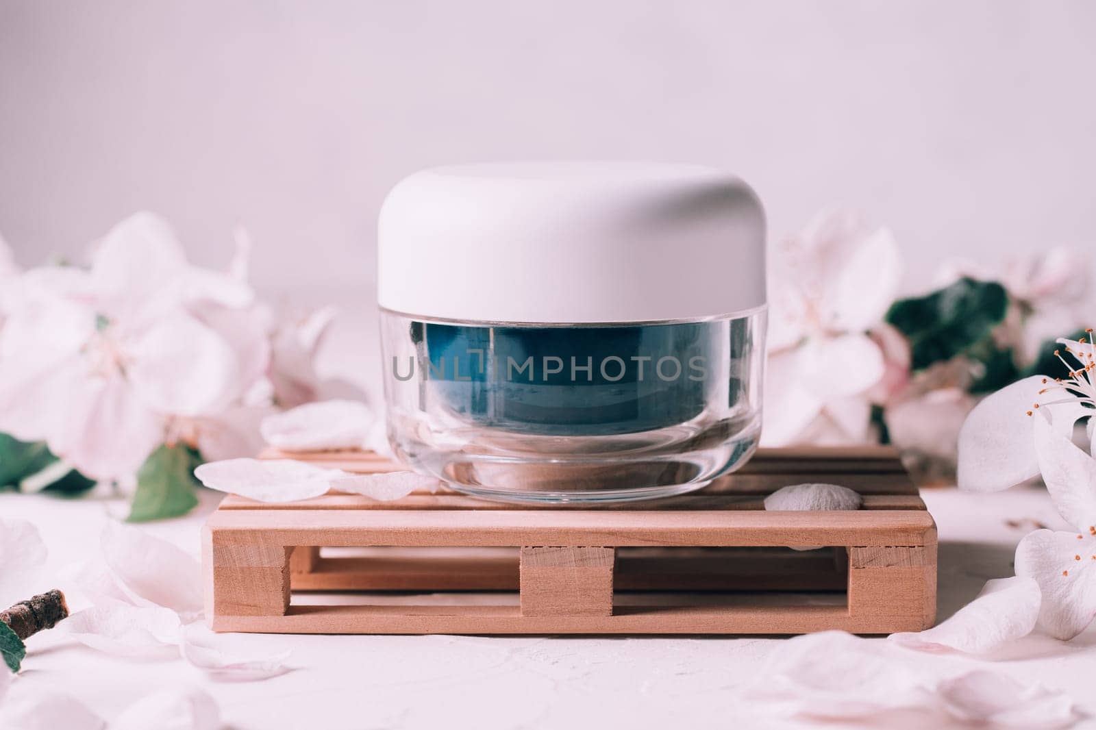 Blue glass jar of cream on wooden podium in form of pallet on light plaster surface with apple flowers