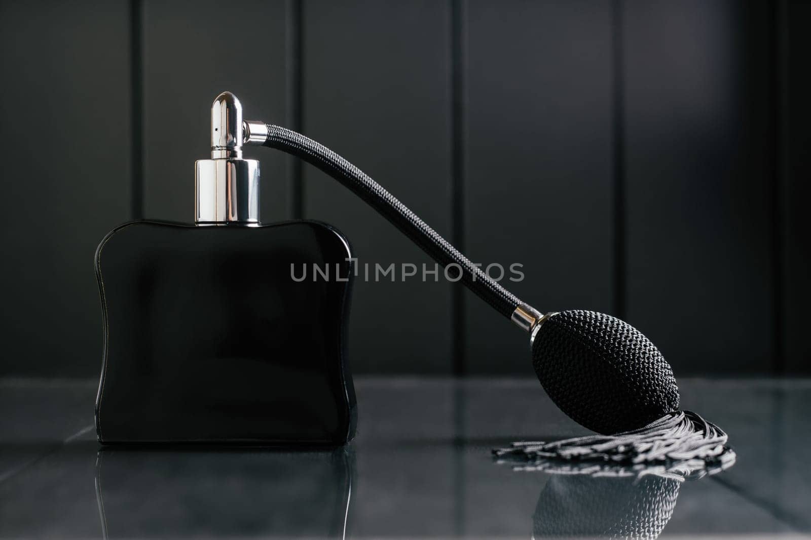 Black bottle of eau de toilette or perfume with long tassel spray pomp stands on dark background on reflective surface. Copy space