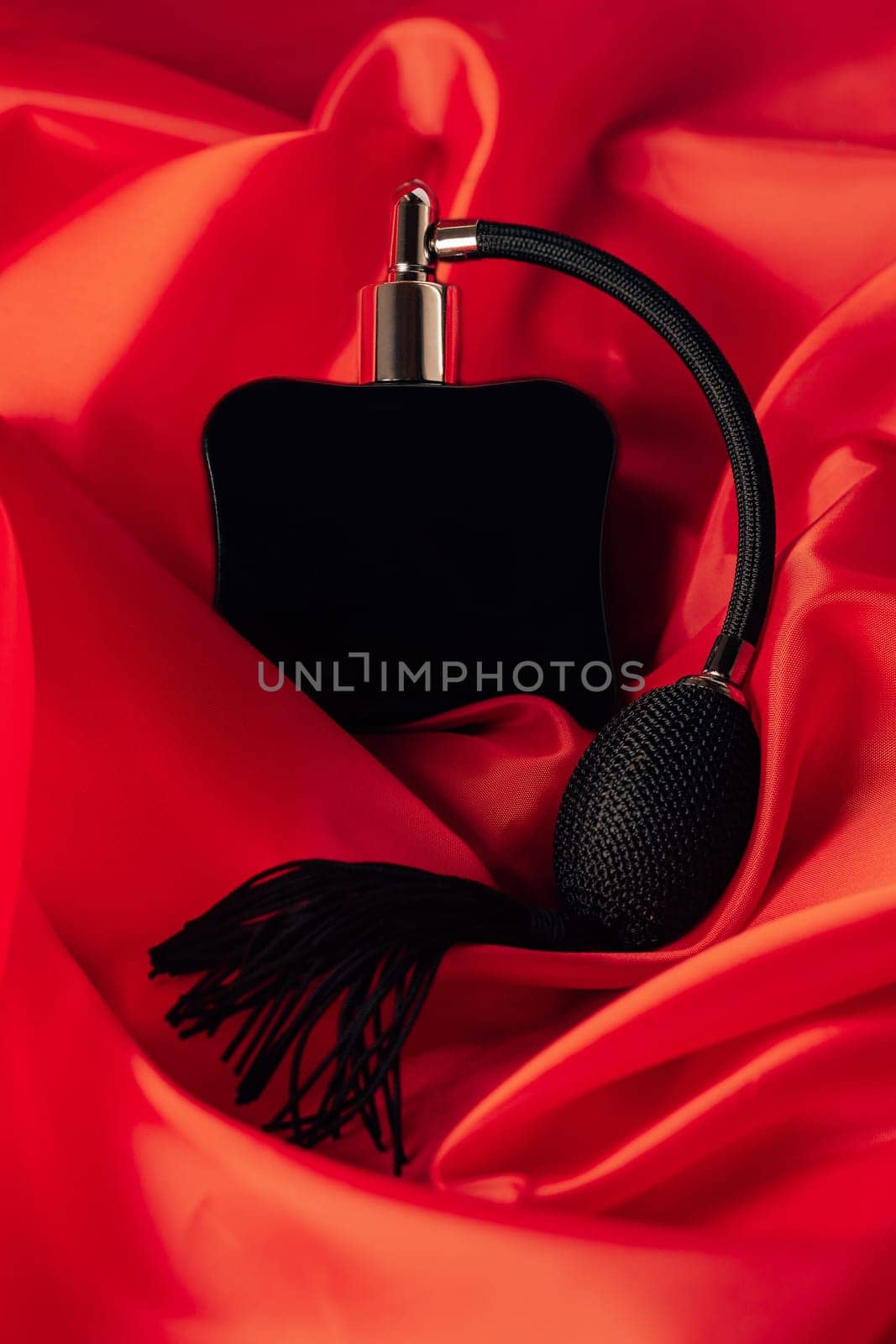 Black bottle of eau de toilette or perfume with long tassel spray pomp lies on red background of flowing fabric. Vertical