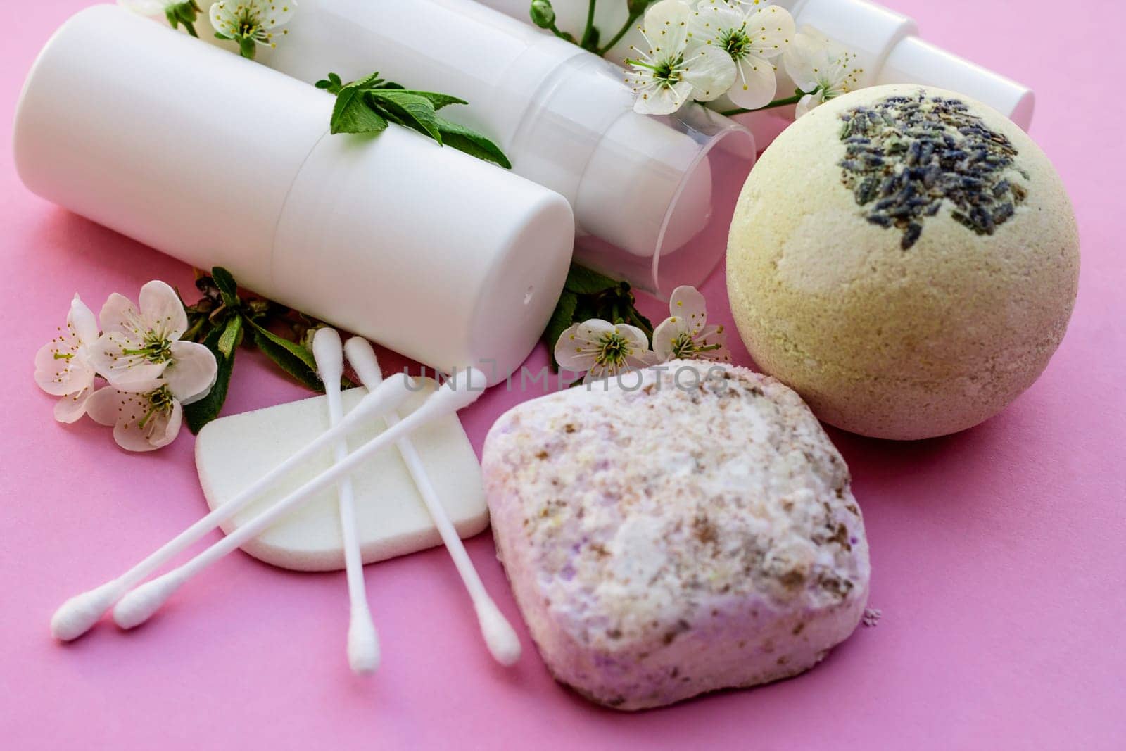 White cosmetic bottles, bath bomb, handmade soap, bath salt, massage brush, sponge, cotton buds with cherry flowers on a pink background. Natural organic cosmetics concept.