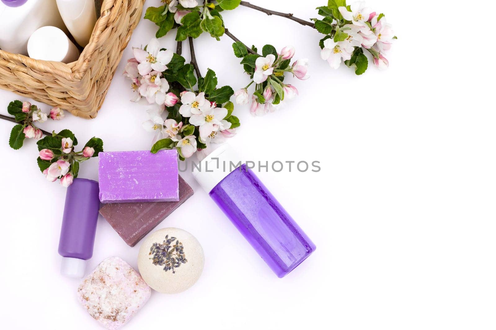 Lilac cosmetic bottles, bath bomb, handmade soap, bath salt near wicker basket with pear flowers on a white background. Natural organic cosmetics concept. Flat lay