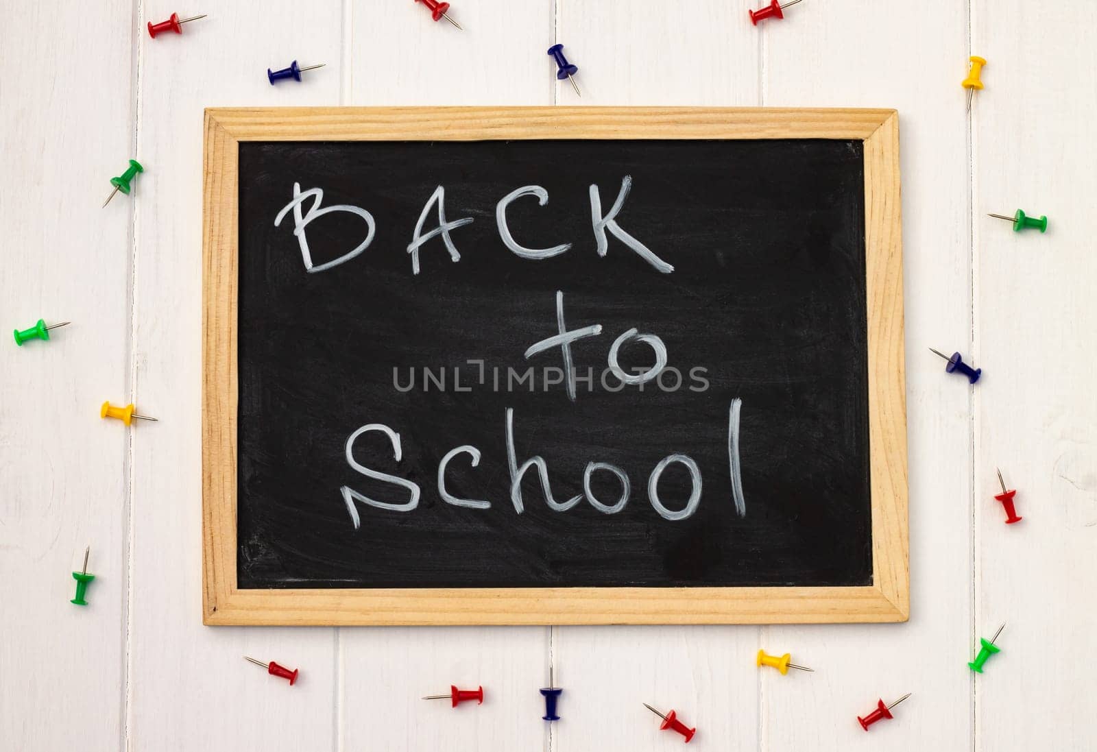 Back to school black chalk board on a white wooden background, color pins. Top view. Copy space. Education concept.