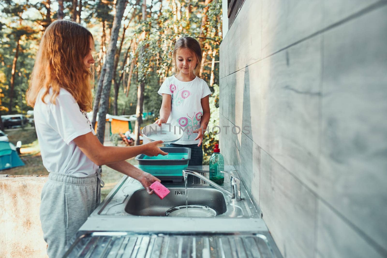 Teenager girl washing up the dishes pots and plates with help her younger sister in the outdoor kitchen during vacations on camping. Camp life. Sisters spending time and working together