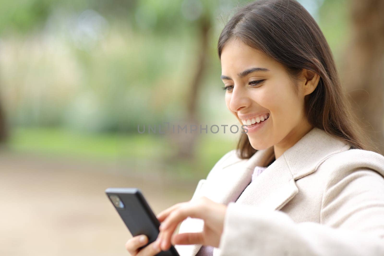 Happy woman in winter using cell phone sitting in a park