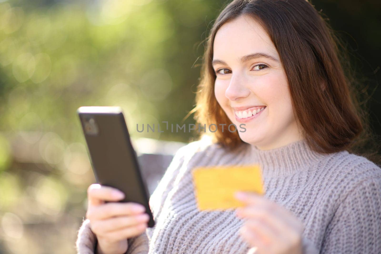 Happy woman looking at you holding cell phone and credit card in a park