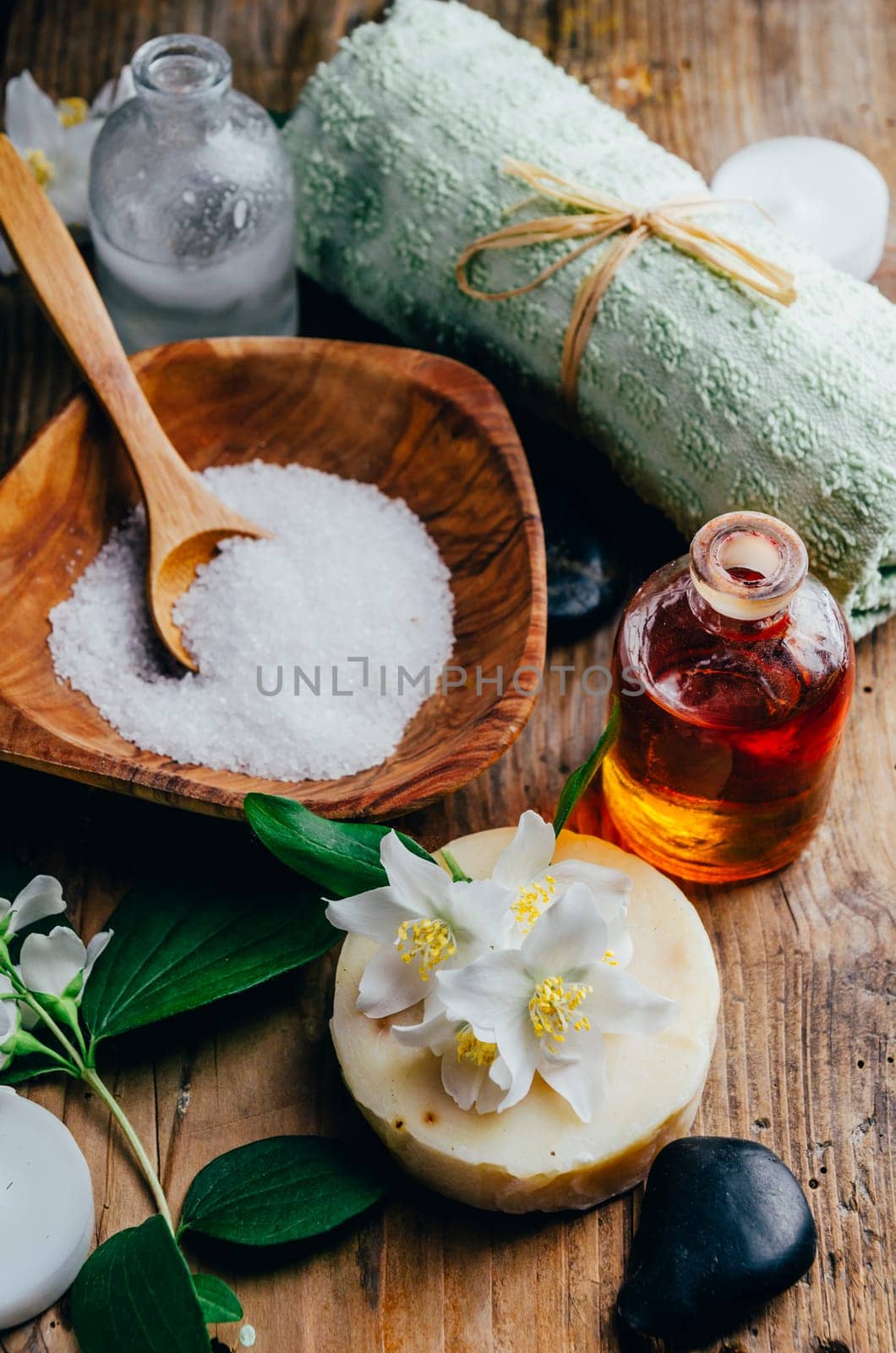 Natural spa cosmetics with essential massage oils, jasmine flower petals, zen stone on rustic wooden background. Handmade soap and herbal aromatherapy wellness treatment. Luxury skincare salon.