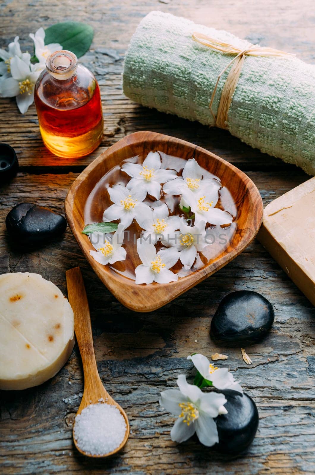 Natural spa cosmetics with essential massage oils, jasmine flower petals, zen stone on rustic wooden background. Handmade soap and herbal aromatherapy wellness treatment. Luxury skincare salon.