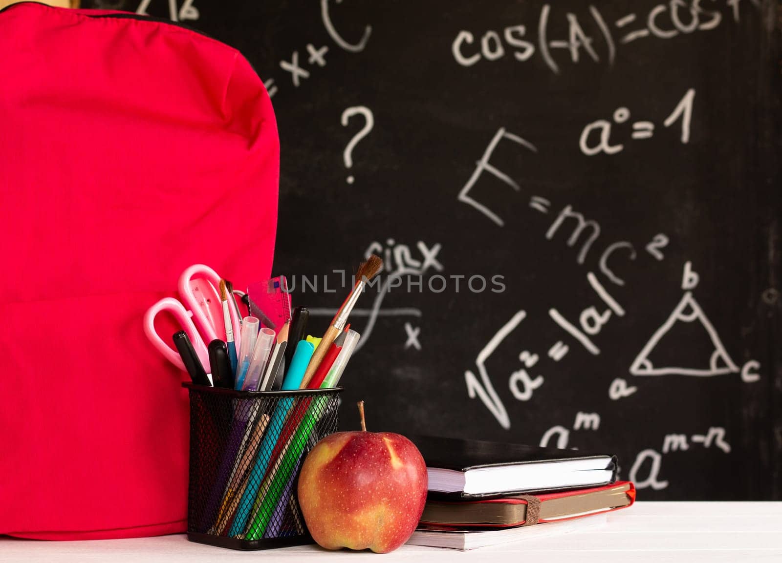 Coloured school supplies, apple, books, backpack on a white contertop on chalkboard background with formulas. Education concept.