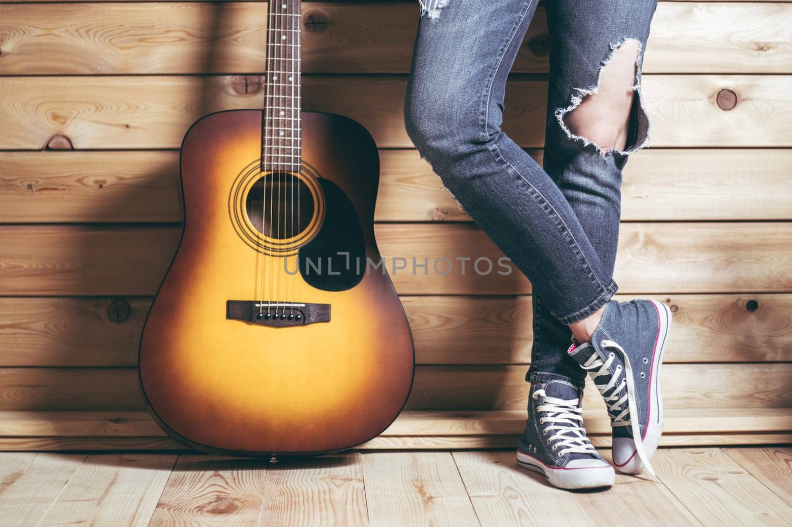 Acoustic six-string yellow-brown guitar and female legs in ripped jeans, near wooden wall