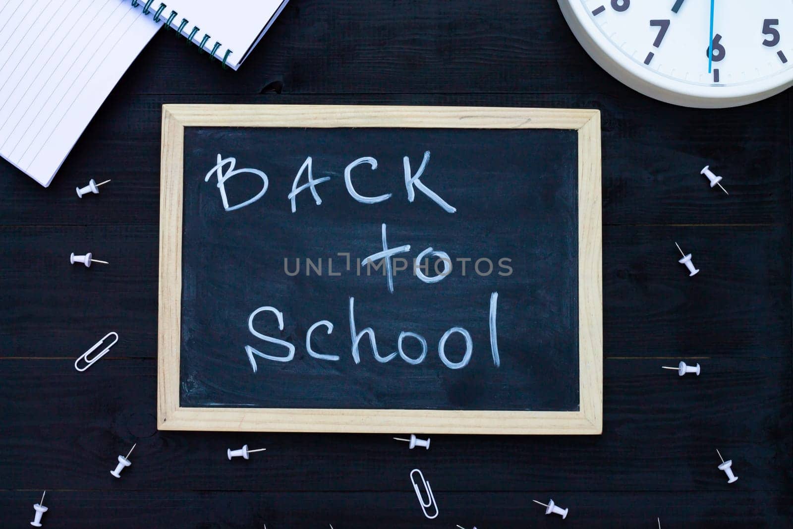 Back to school black chalk board on a black wooden background, white supplies and clock. Top view. Copy space. Education concept.