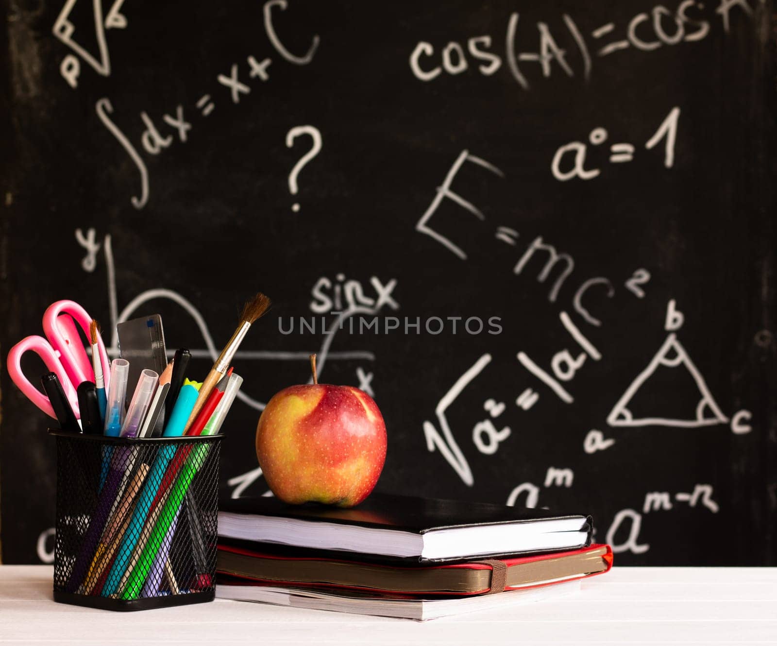 Coloured school supplies, apple, books on a white contertop on chalkboard background with formulas. Education concept