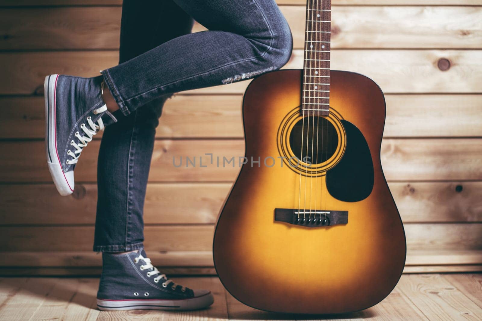 Acoustic six-string yellow-brown guitar and female legs in jeans, near wooden wall