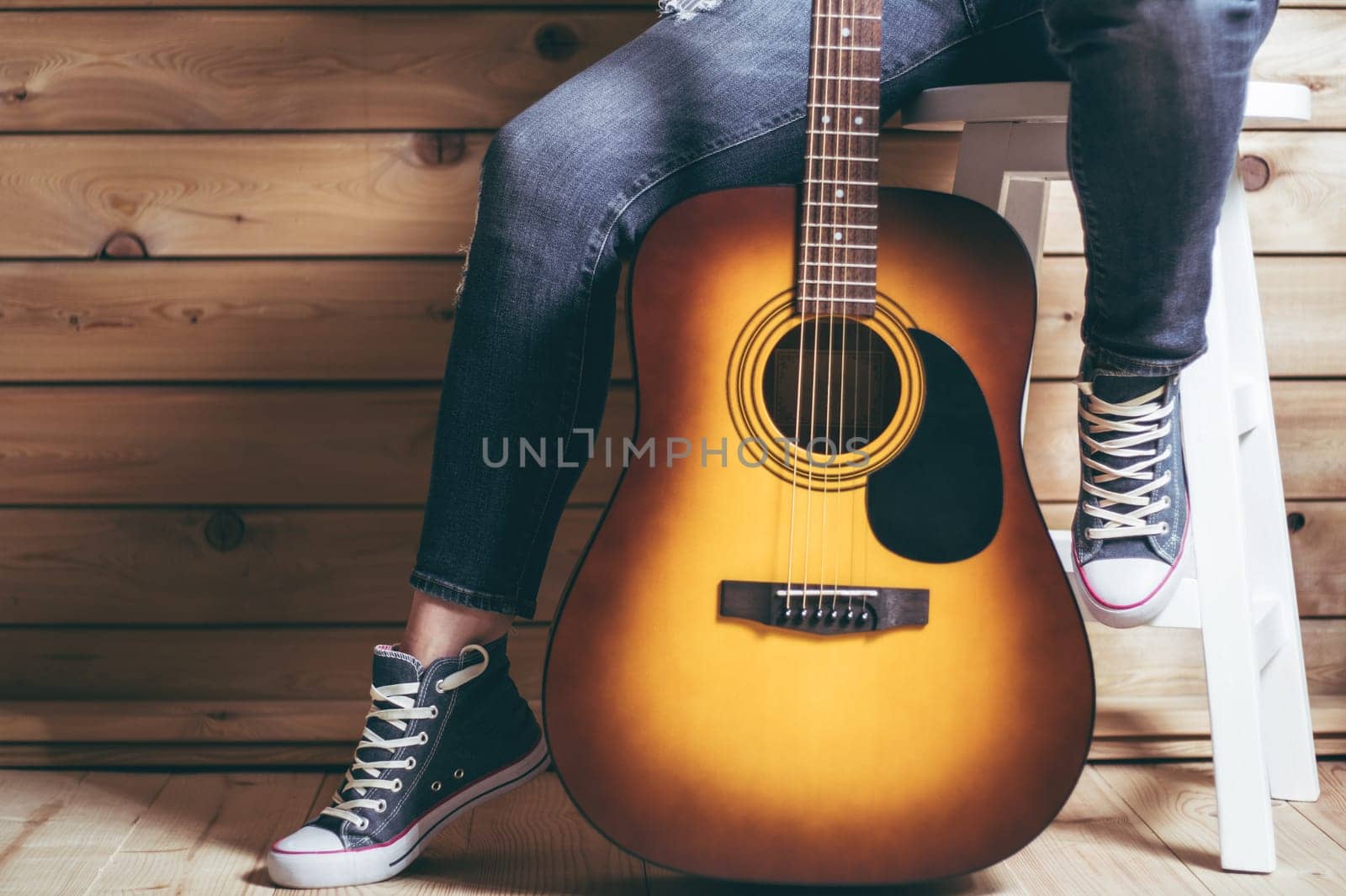 Acoustic six-string yellow-brown guitar and legs of female sitting on stool in jeans, near wooden wall