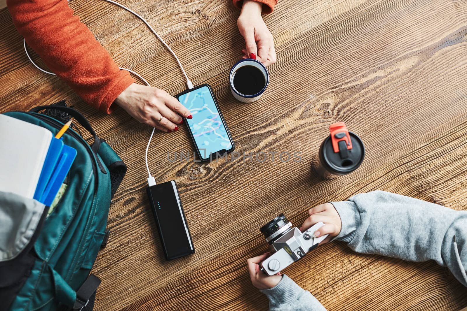 Family planning vacation trip. Woman searching travel destination and routes using navigation map on mobile phone. Girl setting up camera. Charging smartphone with power bank. Using technology while travelling. People sitting at wooden table with camera and backpack during journey. Travel plan concept