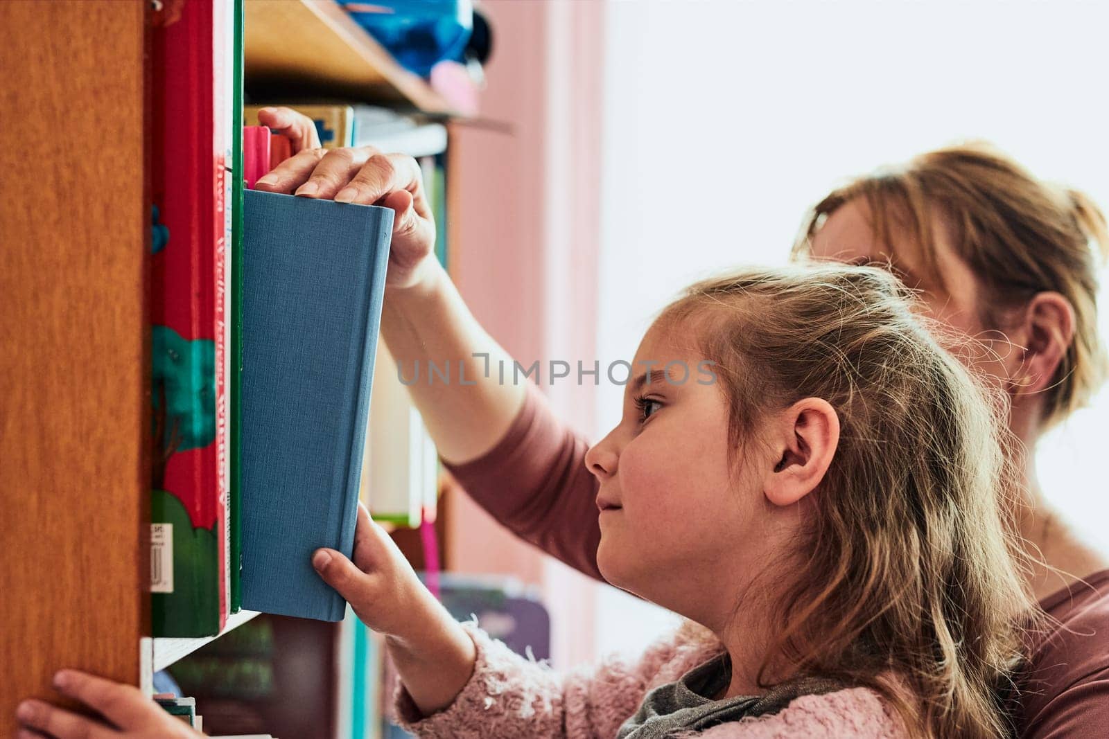 Little girl preschooler choosing a book to read. Child with her mother taking a book from bookshelf