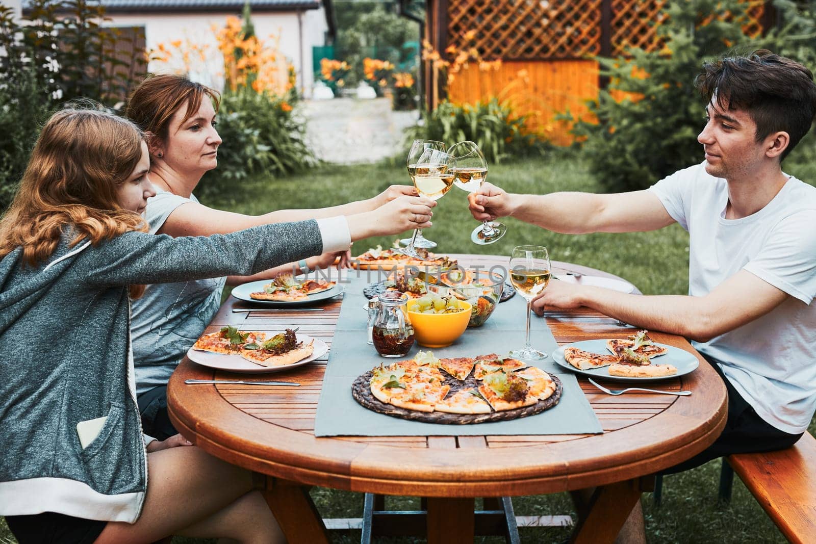 Friends making toast during summer picnic outdoor dinner in a home garden. Close up of people holding wine glasses with white wine over table with pizza, salads and fruits. Dinner in a orchard in a backyard