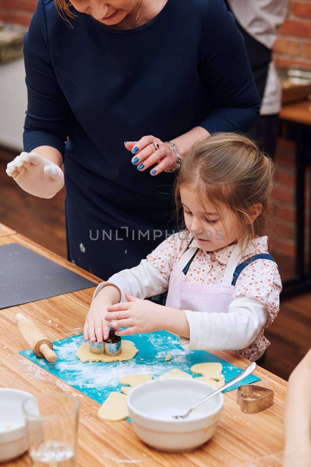 Little girl with her moms help cutting the dough to heart shapes for the cookies. Kids taking part in baking workshop. Baking classes for children, aspiring little chefs. Girls learning to cook. Combining and stirring prepared ingredients. Real people, authentic situations