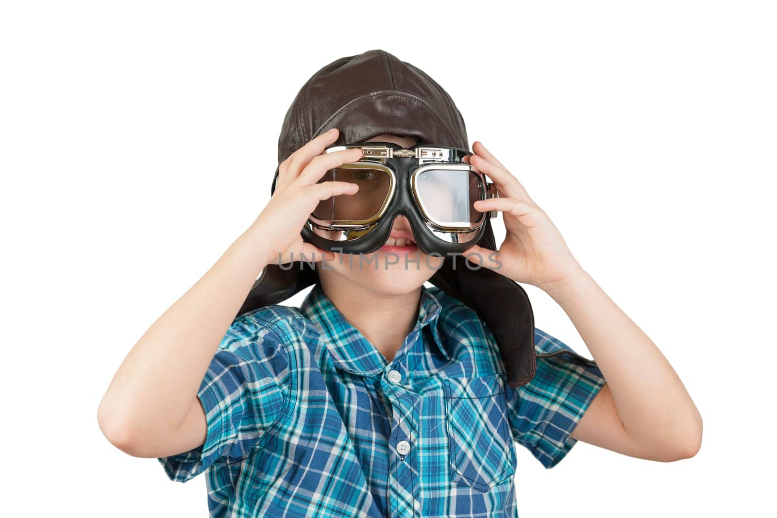 Head and shoulders frontal view of a Caucasian 10 year old boy wearing a leather cap and vintage motorcycle goggles held in front of his face against a white background smiling into the camera.