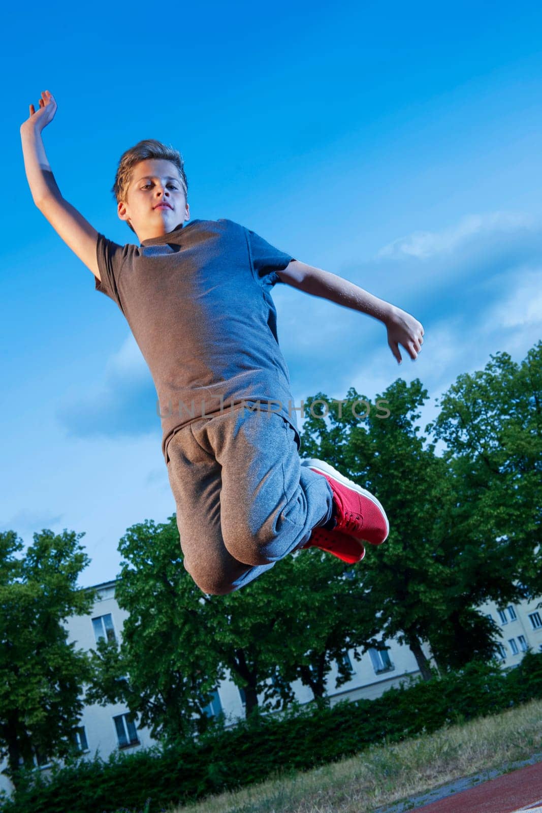 Full body low angle oblique view of teenage boy jumping in the air with arms raised on sports field against blue sky.