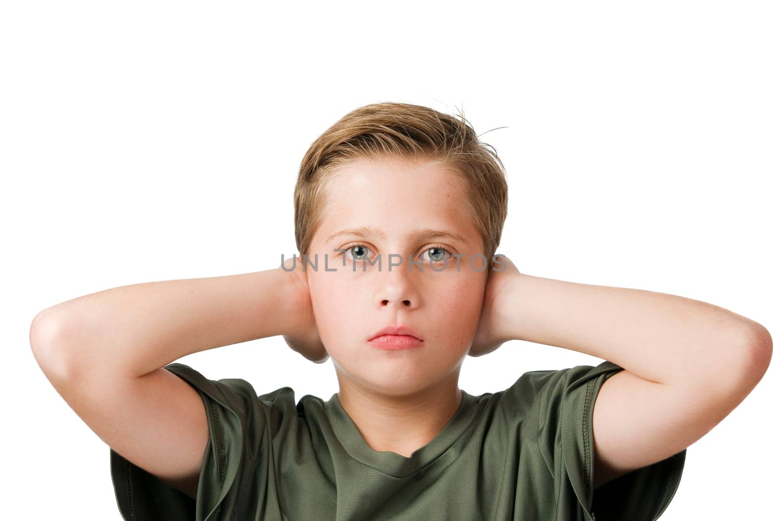 Head and shoulders frontal view of a young male teenage boy in front of a white background covering his ears with his elbows raised and looking seriously at the camera.