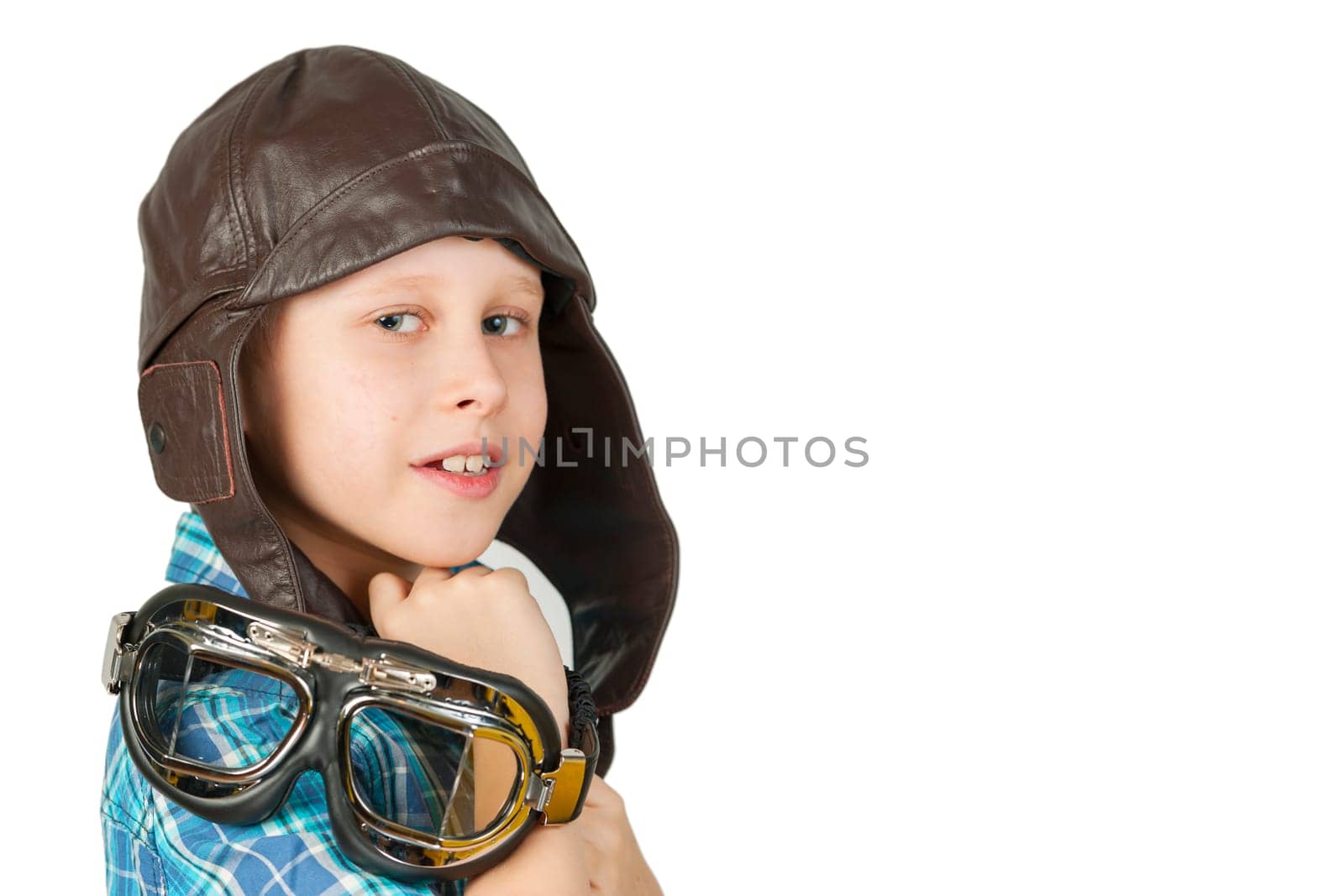 Side head and shoulder view of a 10-year-old Caucasian boy wearing a leather cap and wearing stylish motorcycle goggles over his shoulder against a white background and looking cheekily into the camera.