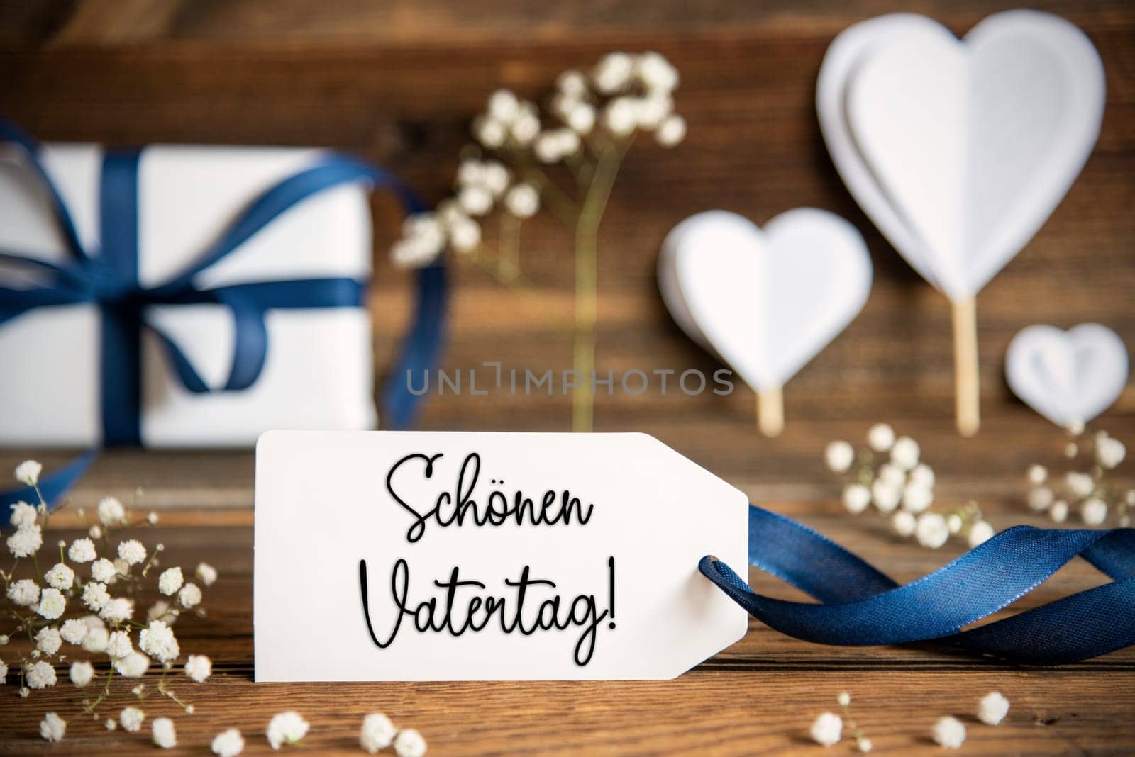 Label With German Text Schoenen Vatertag Means Happy Fathers Day. White Festive Decoration Like Present With Blue Bow, Heart and Flowers. Vintage Wooden Background.