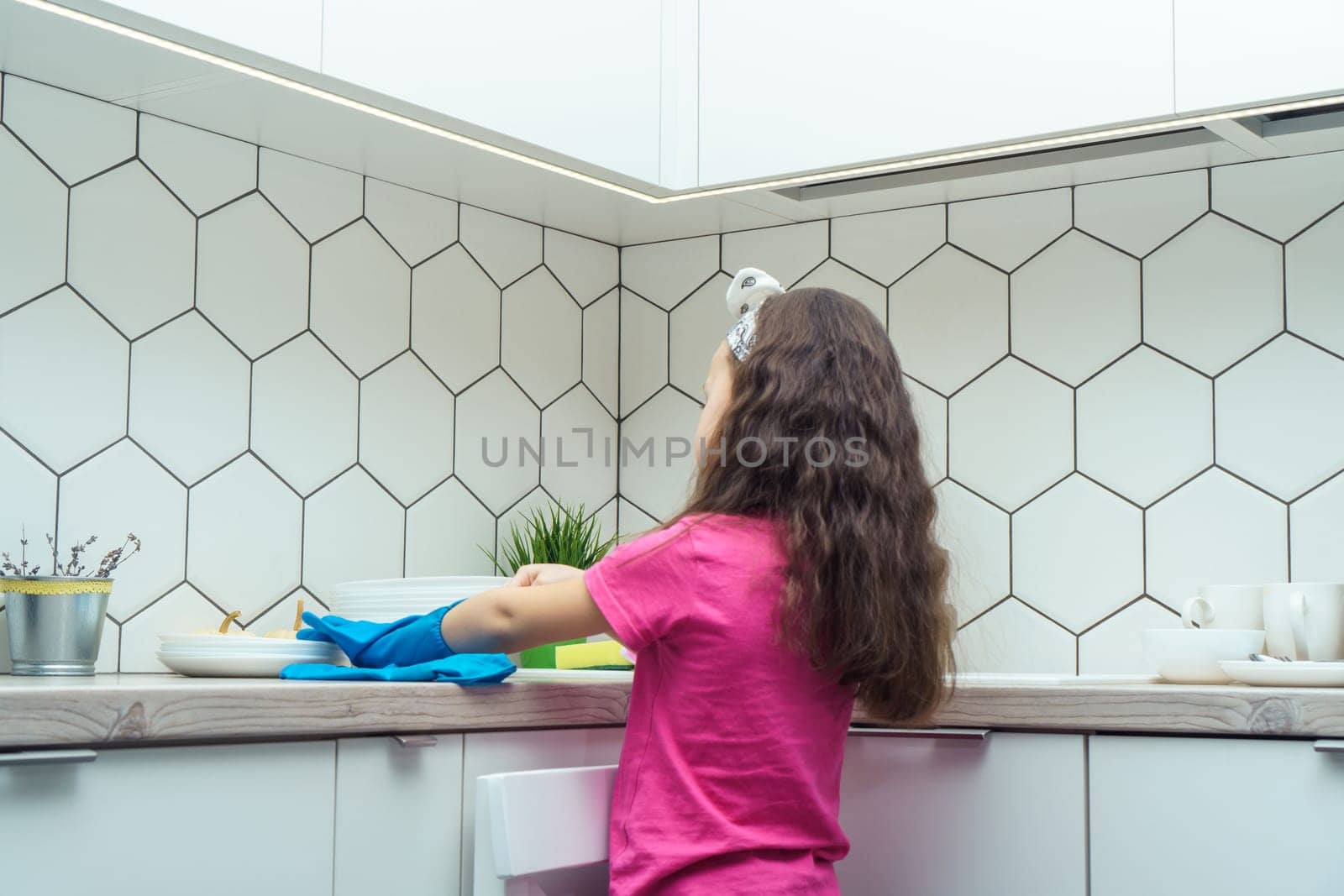 Back view of little preteen girl with loose long dark hair wearing pink T-shirt, taking off blue rubber gloves, standing near table after washing dishes near plates in kitchen. Housework, dishwashing.