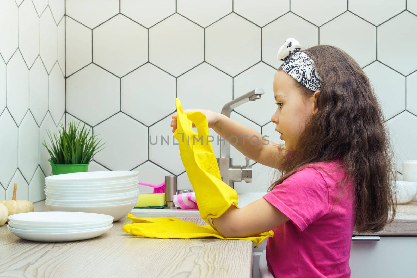 Side view of focused preteen girl with long dark hair wearing pink T-shirt, putting on big yellow rubber gloves, looking at hands, preparing for washing dishes in kitchen. Housework, dishwashing.