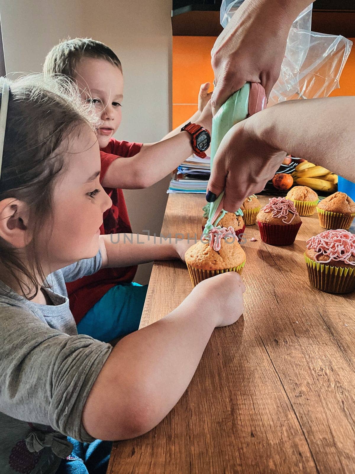Group of children baking cupcakes, squeezing cream from confectionery bag, preparing ingredients, topping, sprinkles for decorating cookies. Kids cooking, working together in kitchen at home. Concept of happy family